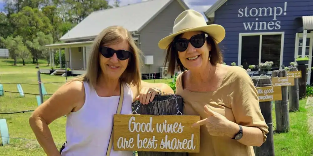 Two middle-aged women, smiling and pointing at a sign at Stomp Wines in the Hunter Valley during a private wine tour, embodying the joy of discovering local wines. The scene invites viewers to book a Hunter Valley wine tasting for a similar experience.