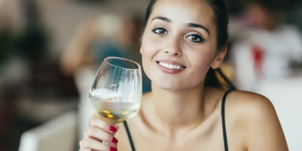 young brunette woman smiling as she enjoys a glass of white wine and displaying wine etiquette