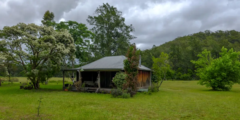 A historic timber hut with a weathered corrugated iron roof stands in the middle of a lightly wooded field in the Mountain View region of the Hunter Valley. Surrounded by tall trees and open grassland, the rustic structure reflects the area's rich heritage. This renowned landscape is home to a vast number of wineries in the Hunter Valley, showcasing the region's renowned winemaking history.