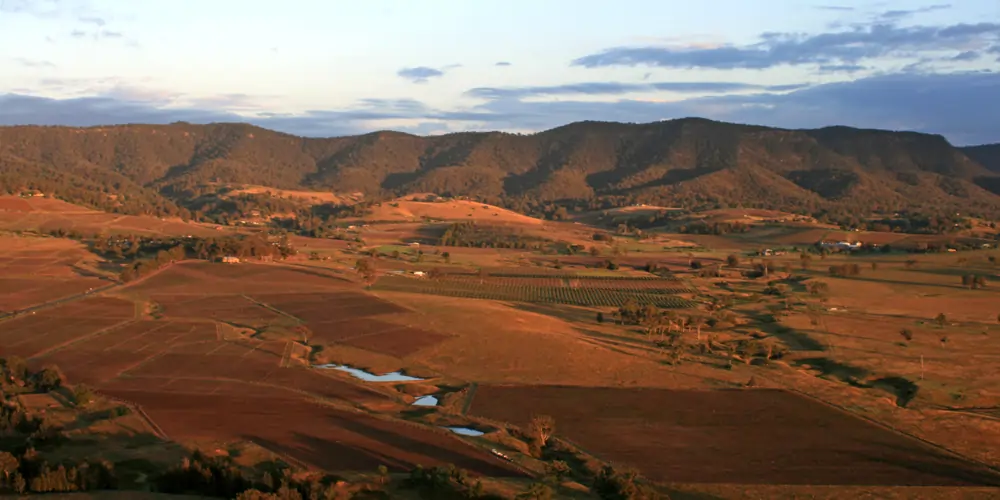 Soft afternoon light washes over the rolling countryside and vineyards of the Hunter Valley in New South Wales, with a blue sky above and clouds tinged purple over a mountain range bathed in golden sunlight — a picturesque view from one of the most historic Australian wine regions