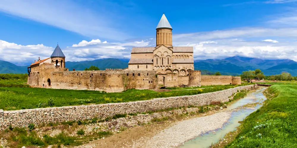 alaverdi-orthodox-monastery-georgia The historic Alaverdi Orthodox Monastery in Georgia, with a stone wall and a stream running alongside it, set against distant mountains, in a region deeply connected to the origin of wine
