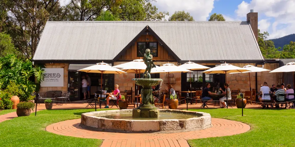 A warm, sunny day lights up the grounds of Café Enzo in the Hunter Valley, where friends and family relax under the shade of umbrellas at timber tables, enjoying glasses of Hunter Valley wine — a lively scene in one of the oldest Australian wine regions