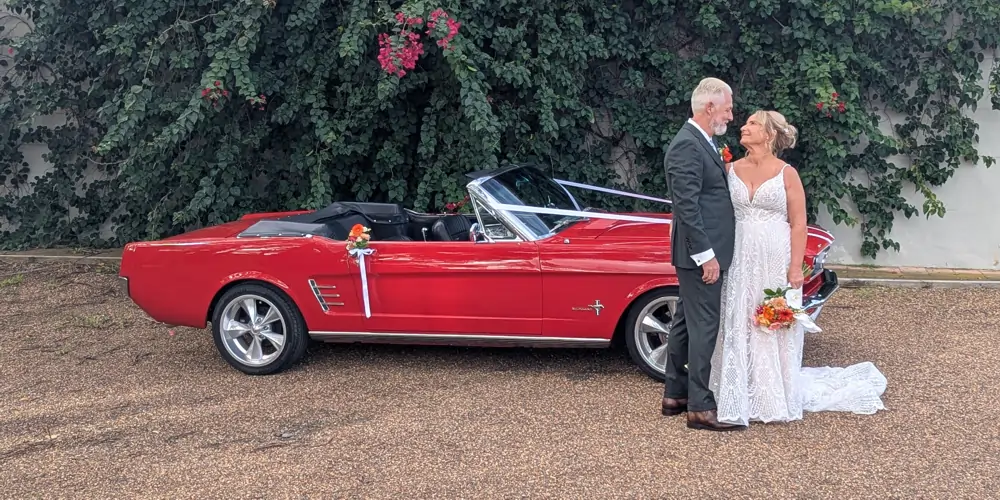 A red 1966 Mustang convertible at a Wandin Estate wedding in the Hunter Valley, with the bride and groom gazing into each other’s eyes in front of the car, a white ribbon on the bonnet and a floral arrangement on the door handle, perfect for Mustang hire