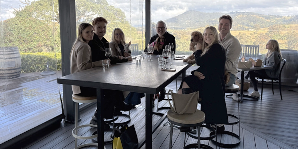 Three couples enjoying a Hunter Valley dog friendly wine tour, smiling with their dogs beside them at a picturesque vineyard with wine glasses in hand and scenic vines in the background